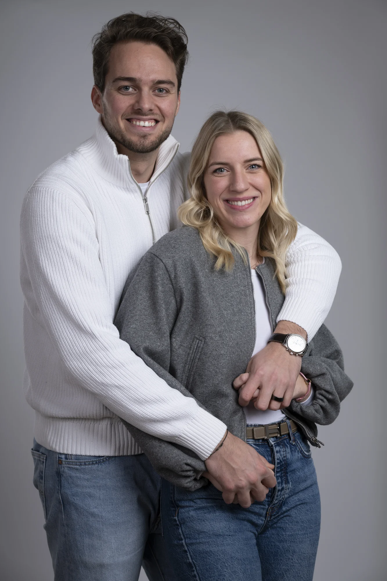 Junges Paar steht eng umschlungen vor grauem Hintergrund, beide lächeln in die Kamera; der Mann trägt einen weißen Pullover, die Frau eine graue Jacke und ein weißes Oberteil.