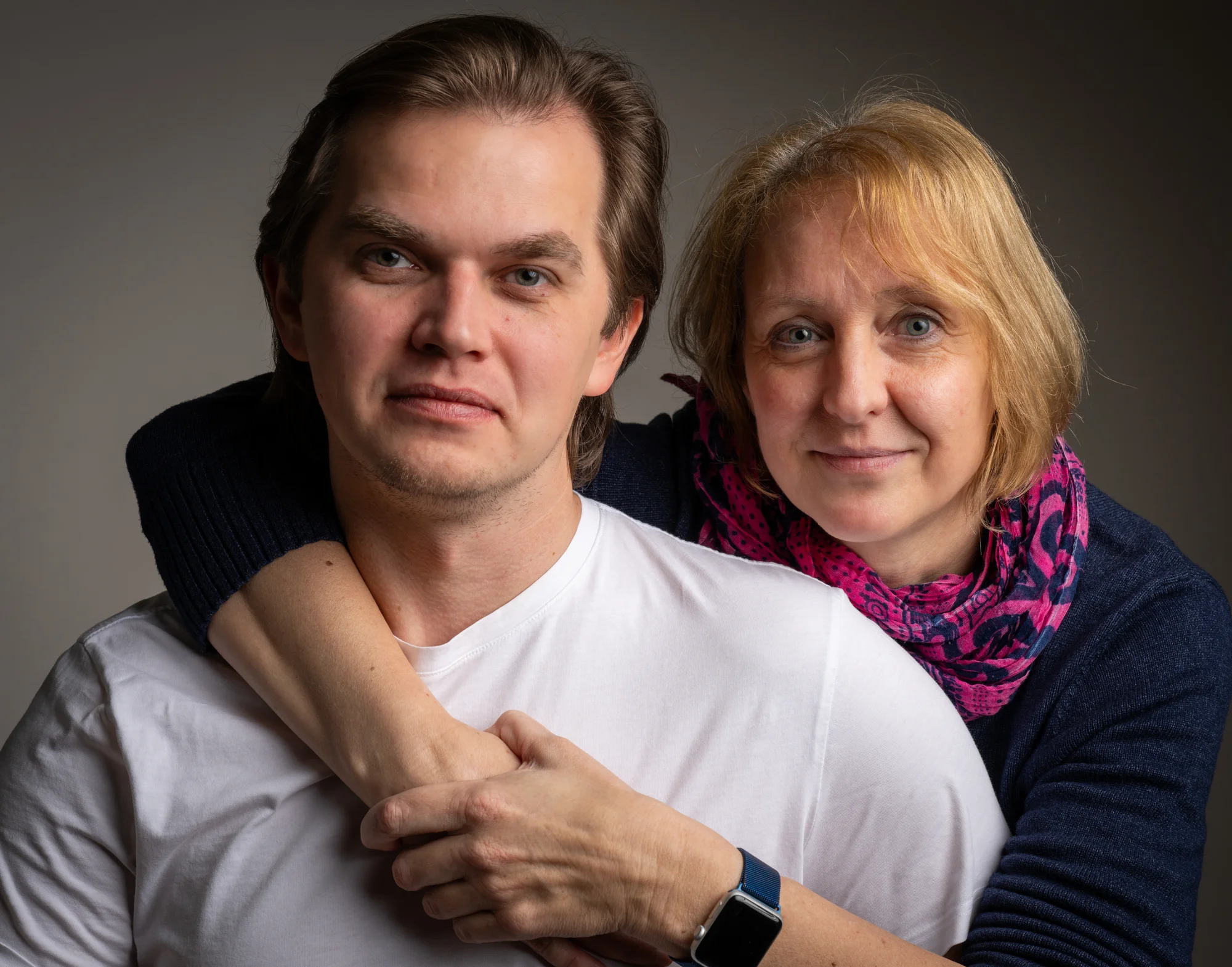 Mittelaltrige Frau umarmt einen jüngeren Mann von hinten, beide blicken ruhig in die Kamera; er trägt ein weißes T-Shirt, sie einen dunkelblauen Pullover mit pink-violettem Schal.