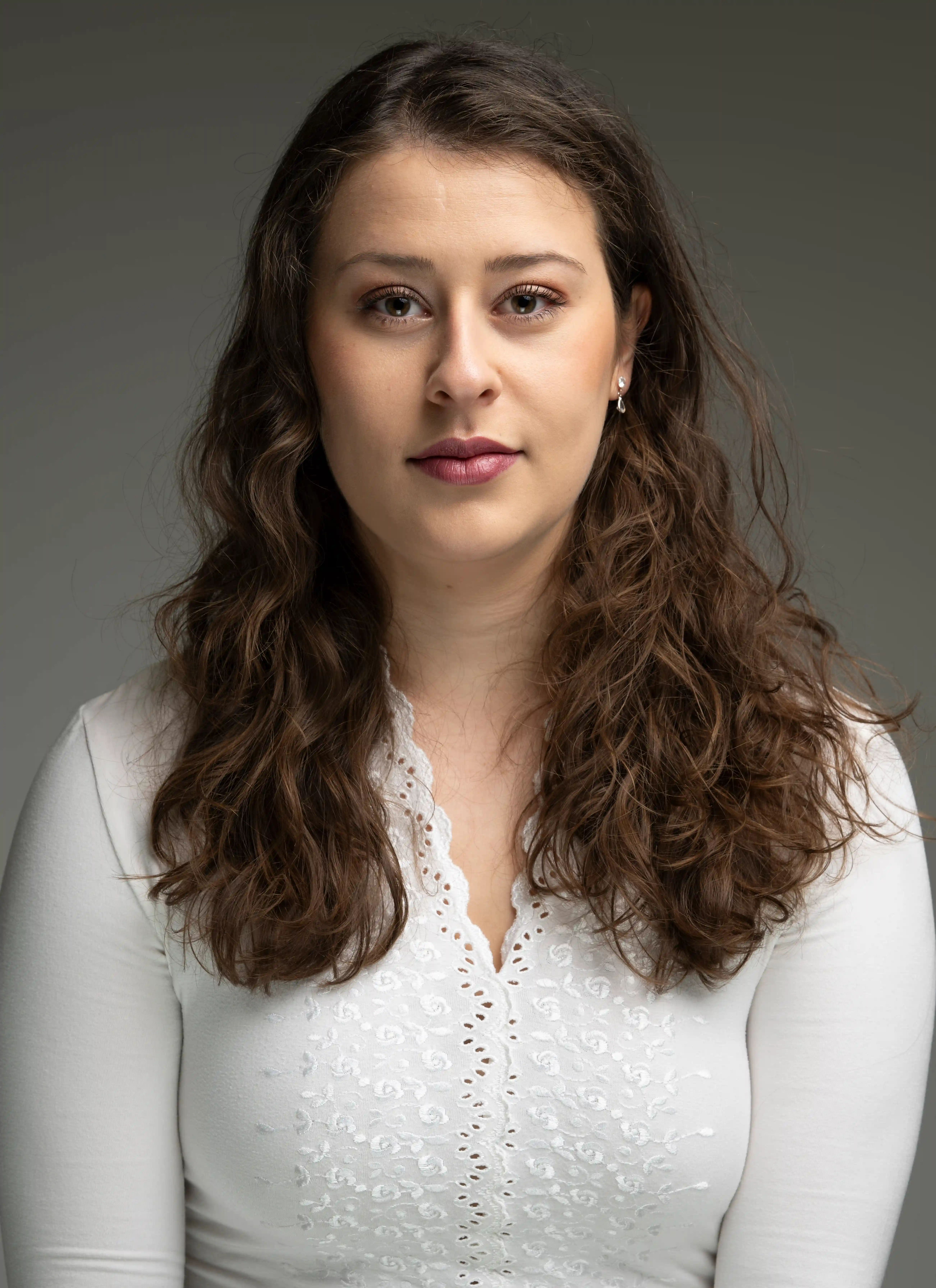 Professionelles Bewerbungsfoto einer jungen Frau mit lockigen braunen Haaren, weißer Bluse und neutralem Hintergrund im Fotostudio.