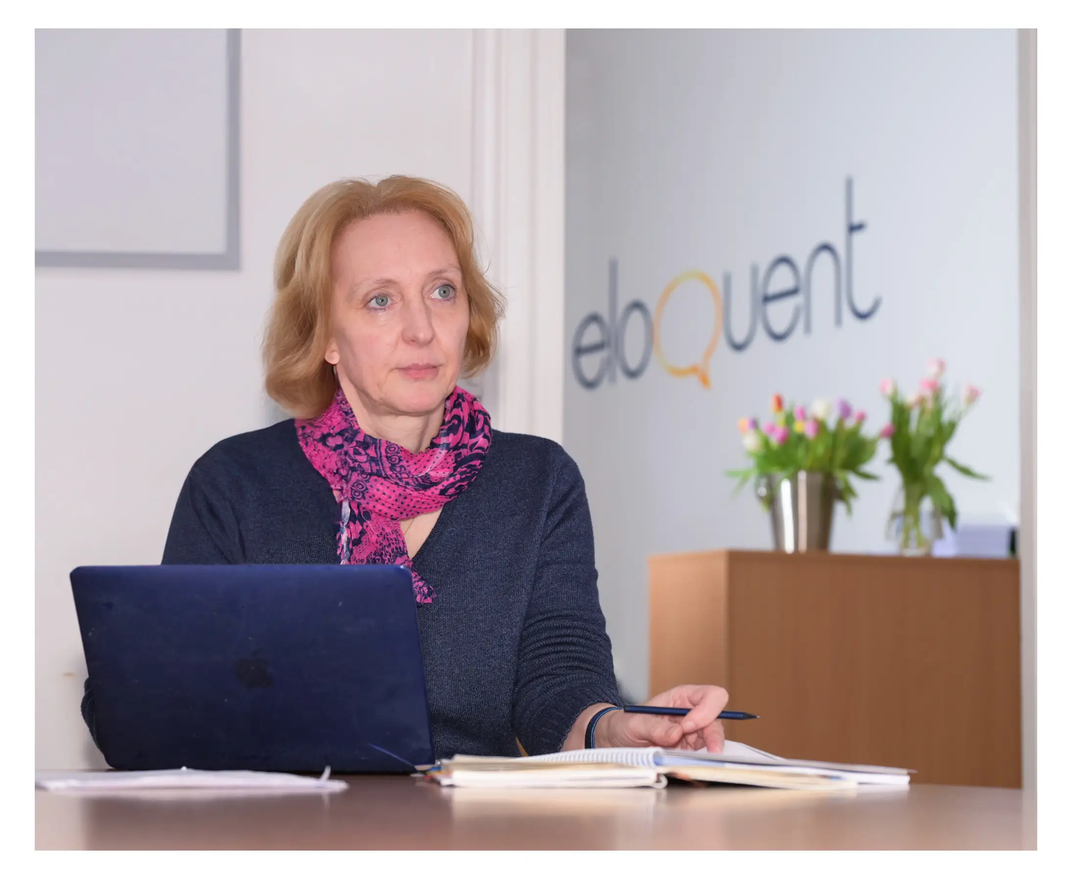 Professionelles Bewerbungsfoto einer Frau mit blonden Haaren im Büro, sitzend vor einem Laptop und Notizbuch, mit pinkem Schal und Firmenlogo im Hintergrund.