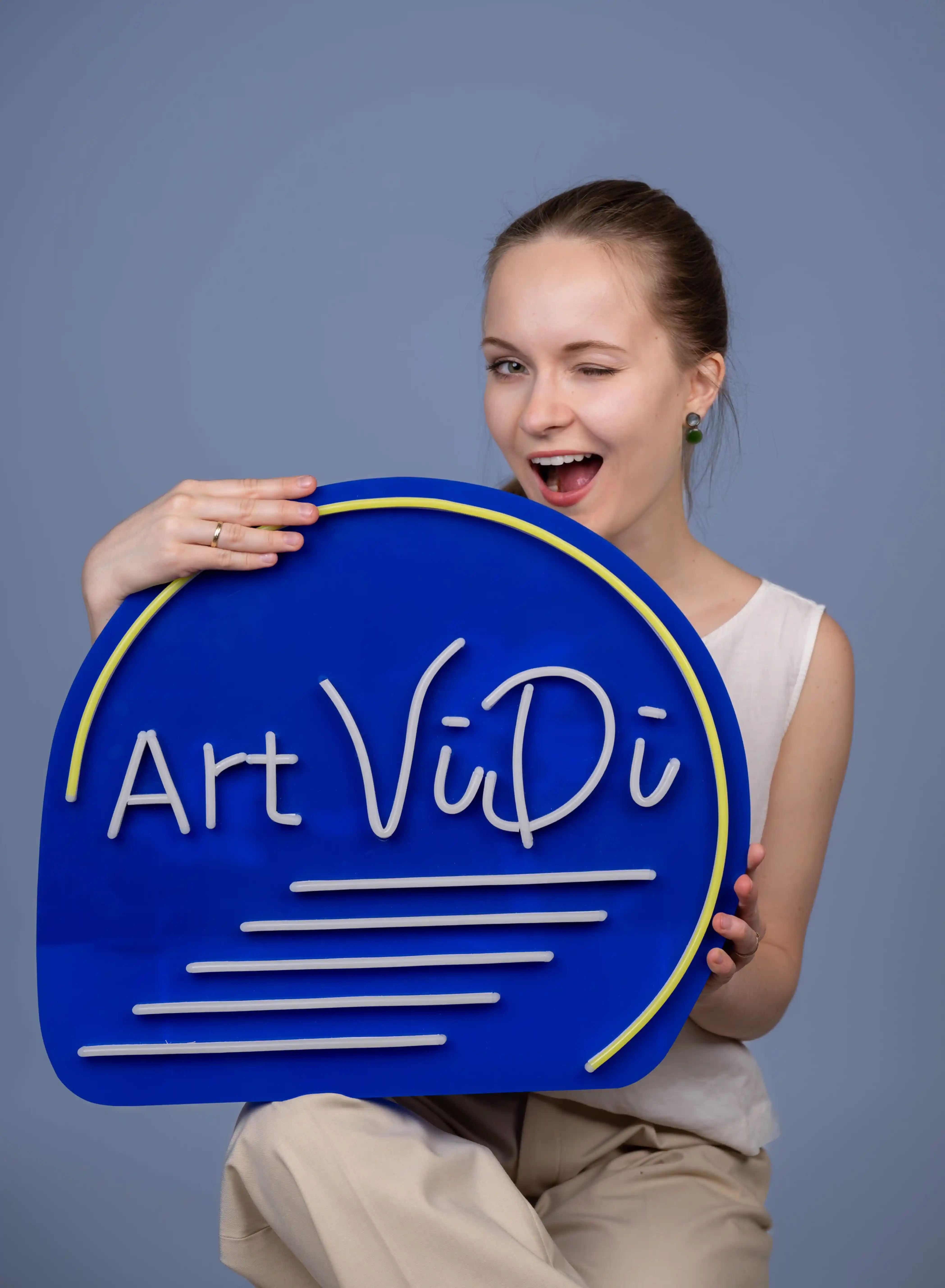 Professionelles Bewerbungsfoto einer jungen Frau in heller Kleidung, die ein blaues Schild mit der Aufschrift „Art ViDi“ hält und dabei zwinkernd in die Kamera lächelt.