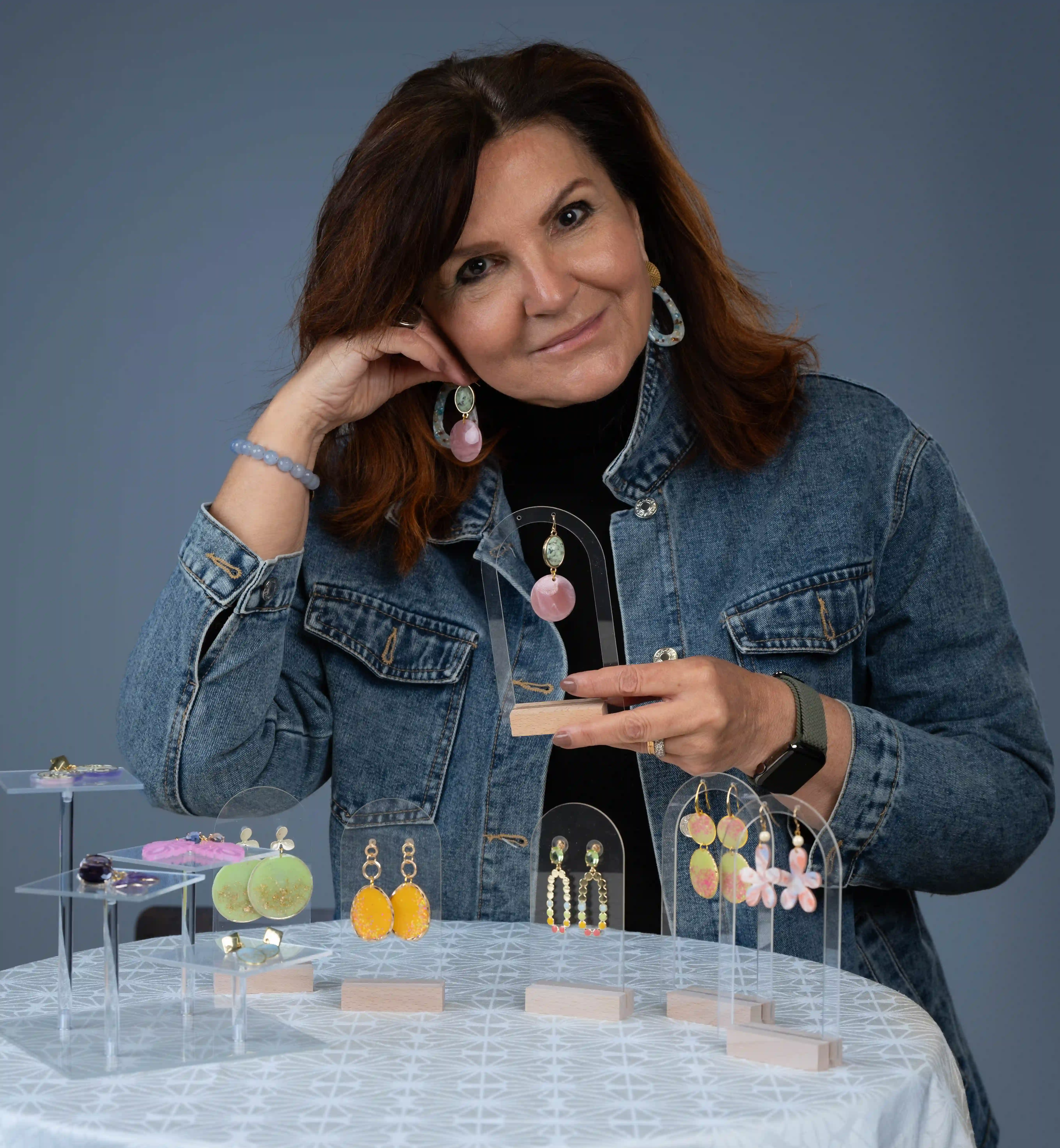 Professionelles Bewerbungsfoto einer Frau mit schulterlangen braunen Haaren in Jeansjacke, die farbenfrohe Ohrringe präsentiert, aufgenommen vor blauem Studiohintergrund.