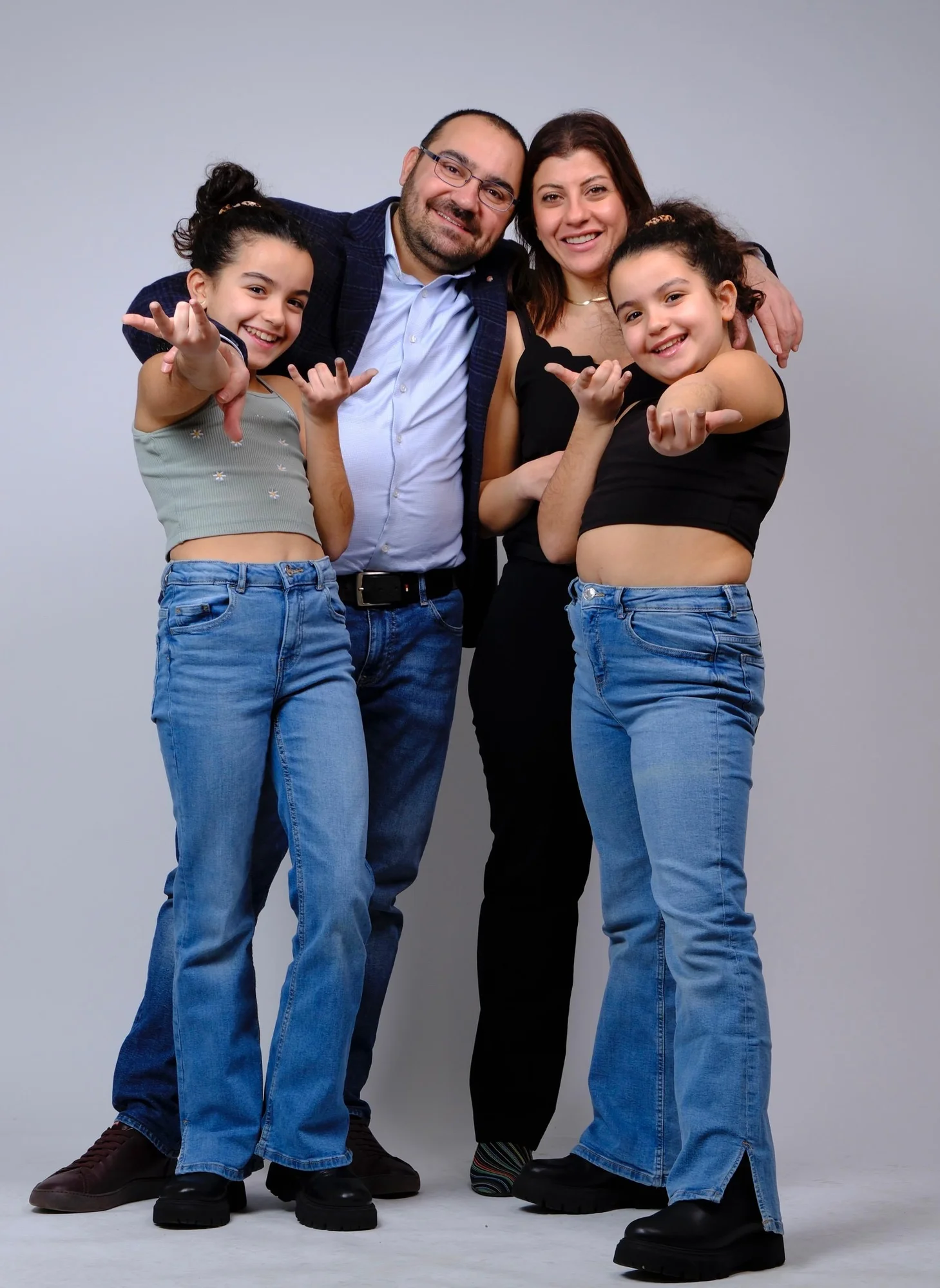 Familie mit Vater, Mutter und zwei Zwillingsmädchen, alle lächelnd und in lockerer Pose, vor neutralem Studiohintergrund.