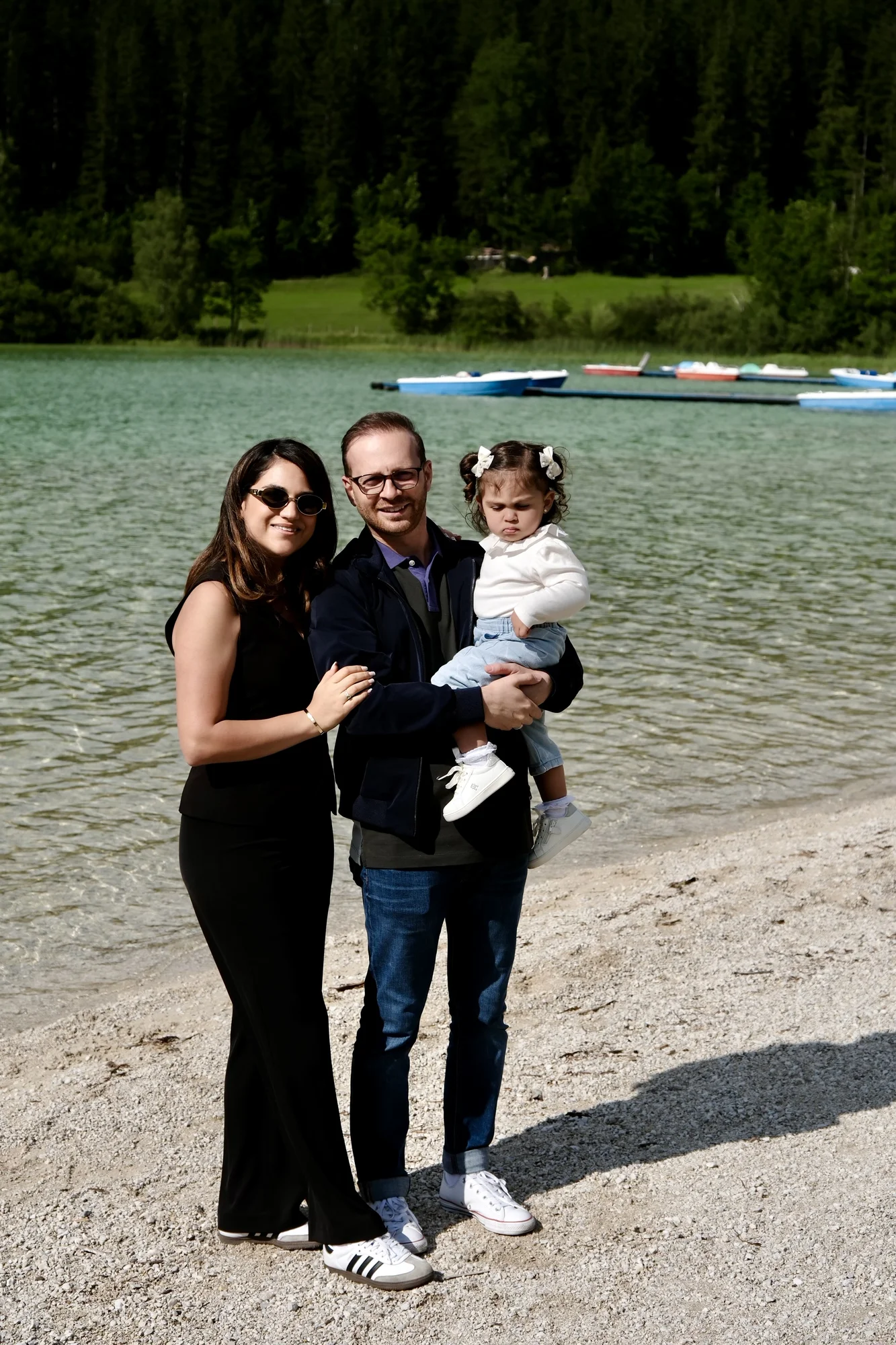 Familienfoto am Seeufer mit einer Frau in schwarzem Outfit, einem Mann in Jeans und dunkler Jacke sowie einem kleinen Mädchen im weißen Pullover, vor türkisfarbenem Wasser und Wald im Hintergrund.