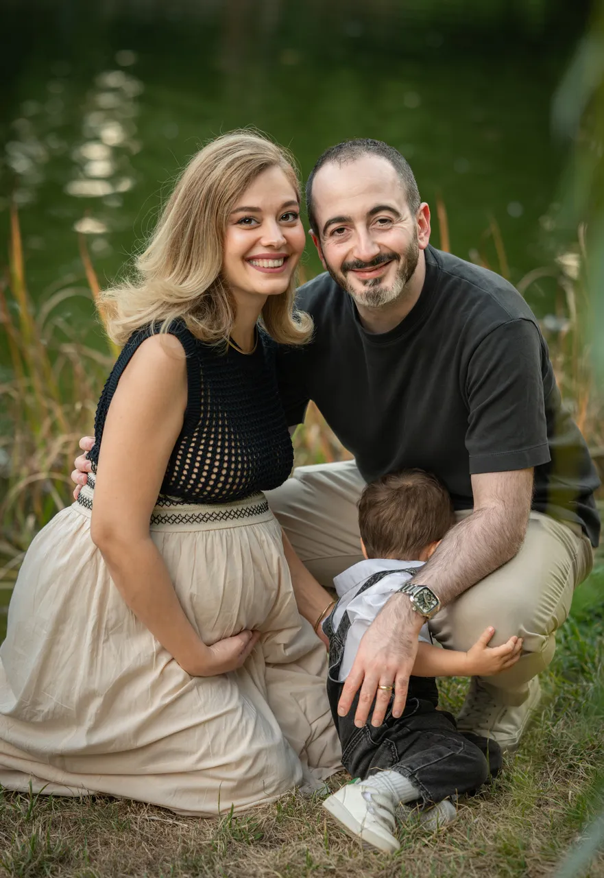 Familienfoto am Seeufer mit einer Frau in schwarzem Outfit, einem Mann in Jeans und dunkler Jacke sowie einem kleinen Mädchen im weißen Pullover, vor türkisfarbenem Wasser und Wald im Hintergrund.