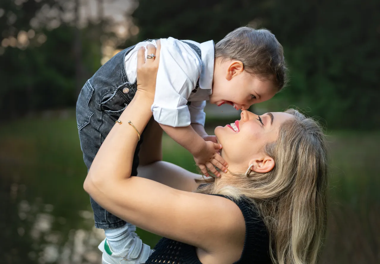 Familienfoto am Seeufer mit einer Frau in schwarzem Outfit, einem Mann in Jeans und dunkler Jacke sowie einem kleinen Mädchen im weißen Pullover, vor türkisfarbenem Wasser und Wald im Hintergrund.